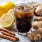 Glas mit Ingwer‑Zimt‑Cola, umgeben von frischem Ingwer, Zimtstangen und Zitrone